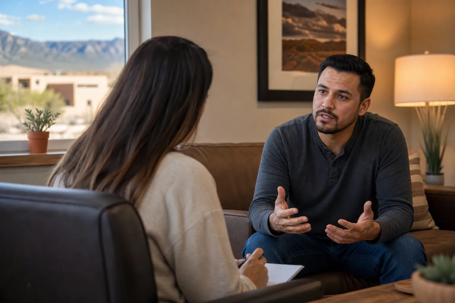A man gestures while speaking with a woman holding a pen and notepad in a warmly lit office with mountain views.