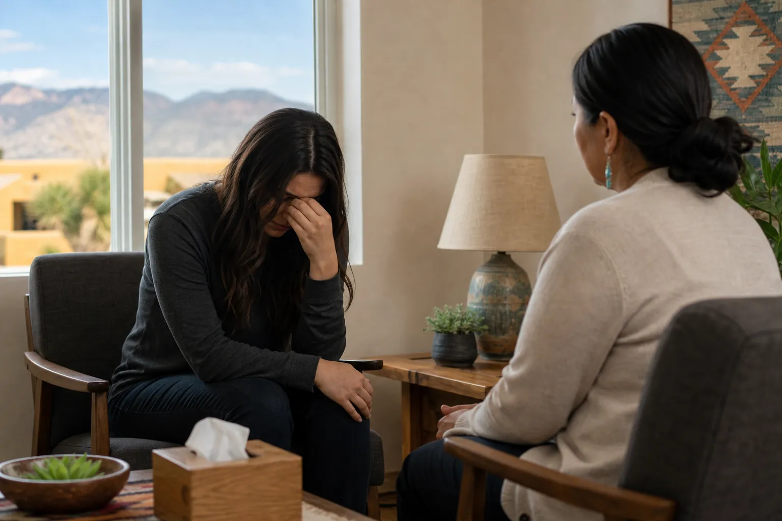 A distressed woman covers her face while sitting opposite another woman in a counseling room with tissues and mountain views.