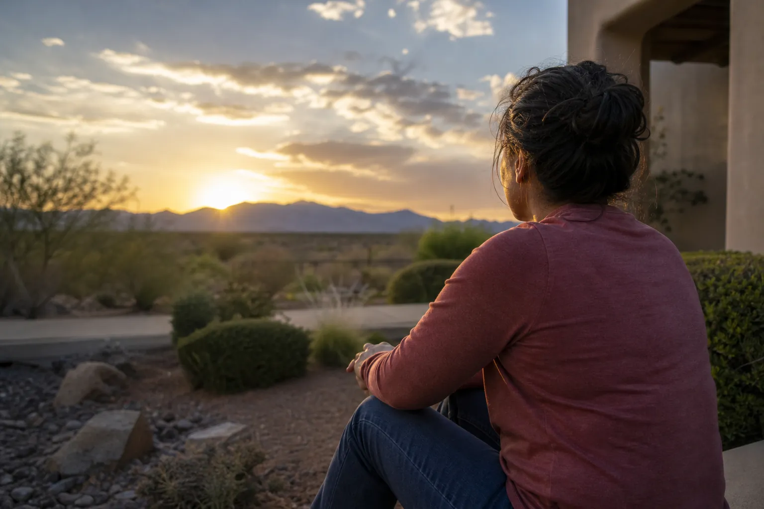A woman in a pink long-sleeved shirt and jeans sits outdoors facing a sunset over desert shrubs and distant mountains.