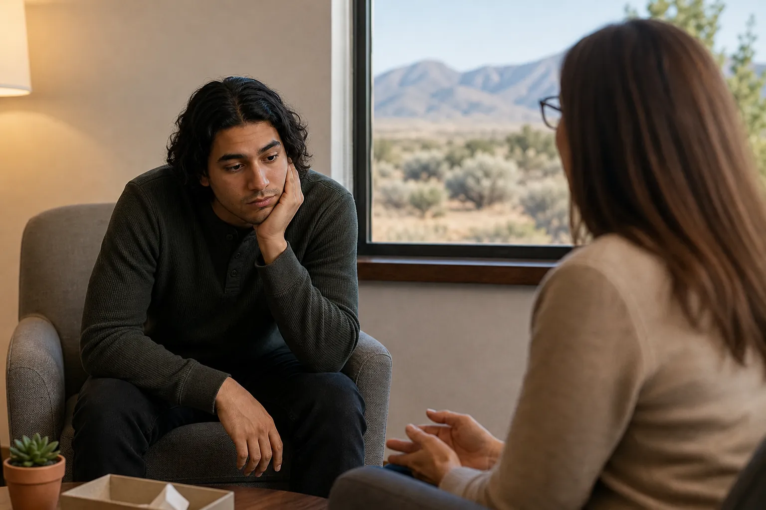 A man rests his cheek on his hand while a woman gestures opposite him, with desert mountains visible through a window.