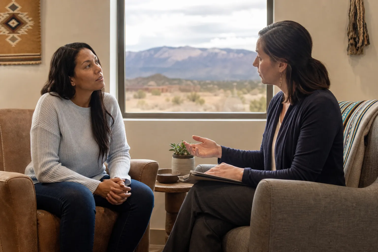 Two women sit facing each other in armchairs, one gesturing with a notebook on her lap, beside a window overlooking mountains.