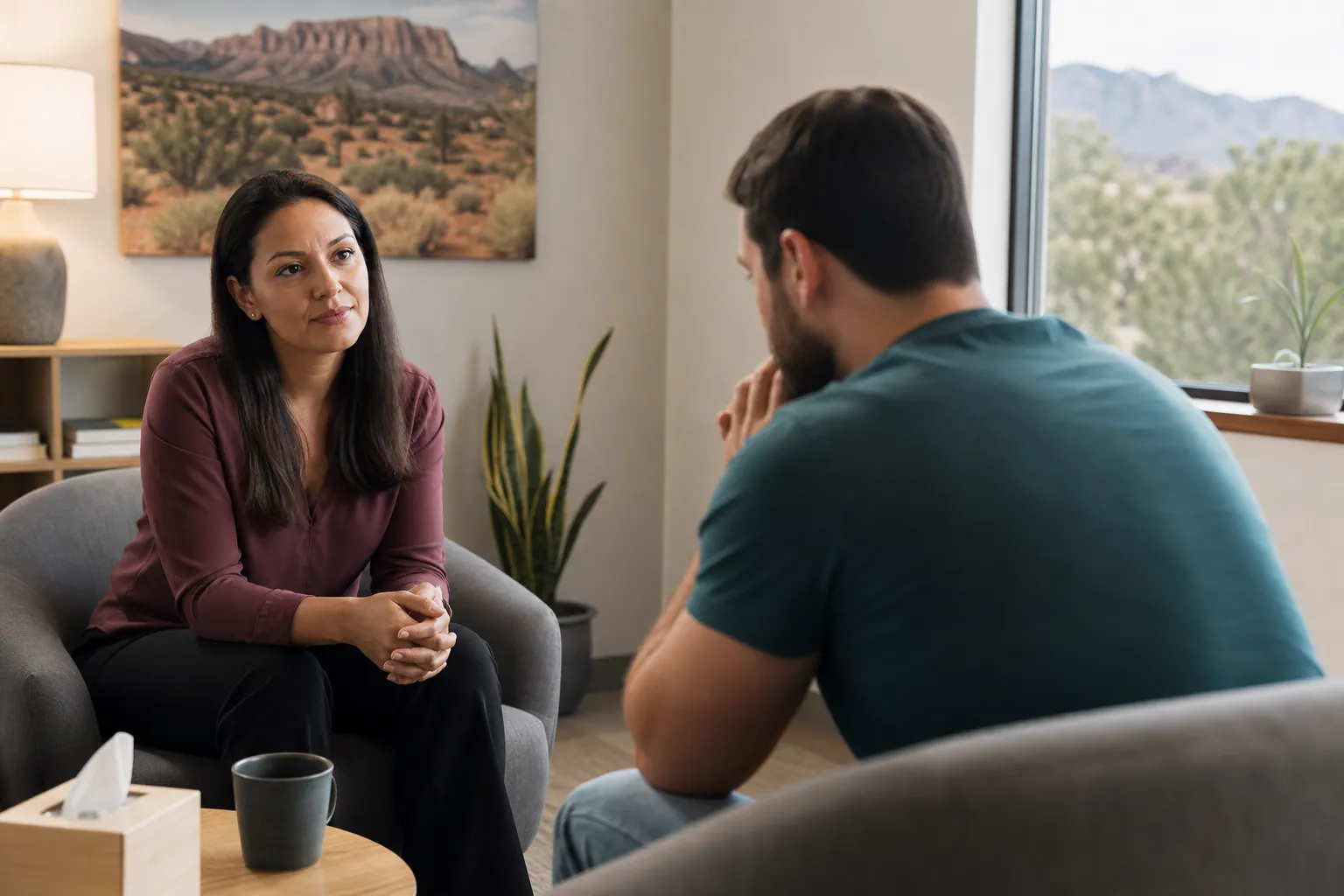 A woman listens to a man seated opposite her in a counseling office with desert artwork, plants, and a tissue box.