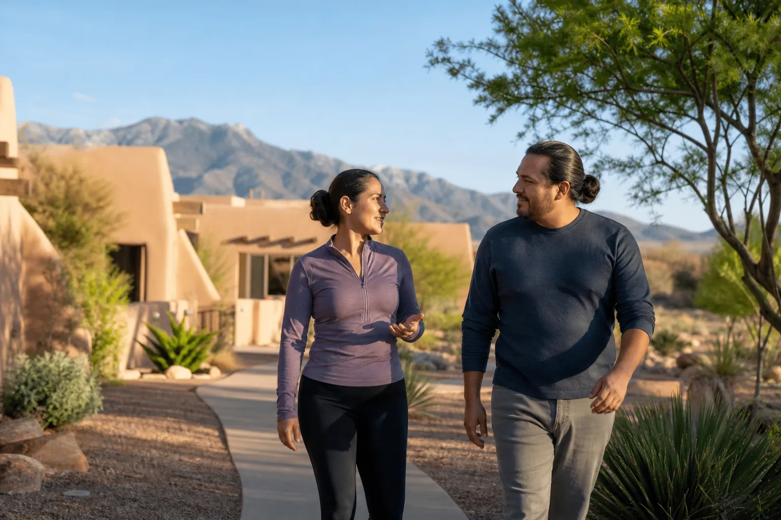 A woman and man walk and talk on a landscaped path beside adobe buildings, with desert plants and mountains behind them.