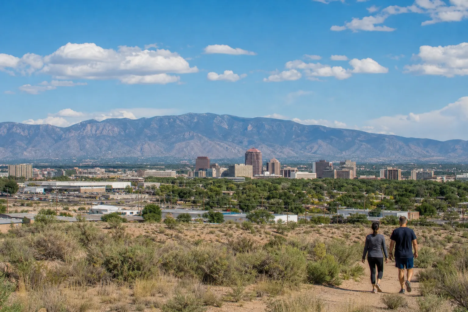Two people walk along a dry, shrub-lined trail overlooking Albuquerque's skyline and mountains beneath a partly cloudy blue sky.