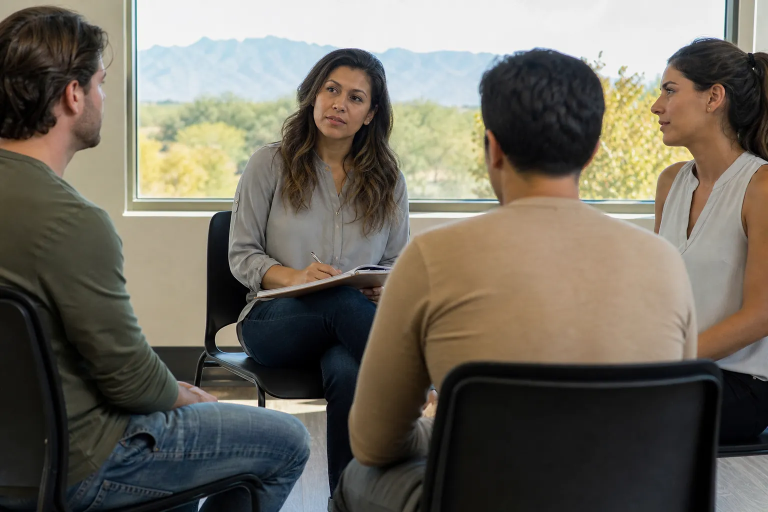 A woman holding a pen and notebook sits with three adults in a counseling circle beside a window overlooking mountains.