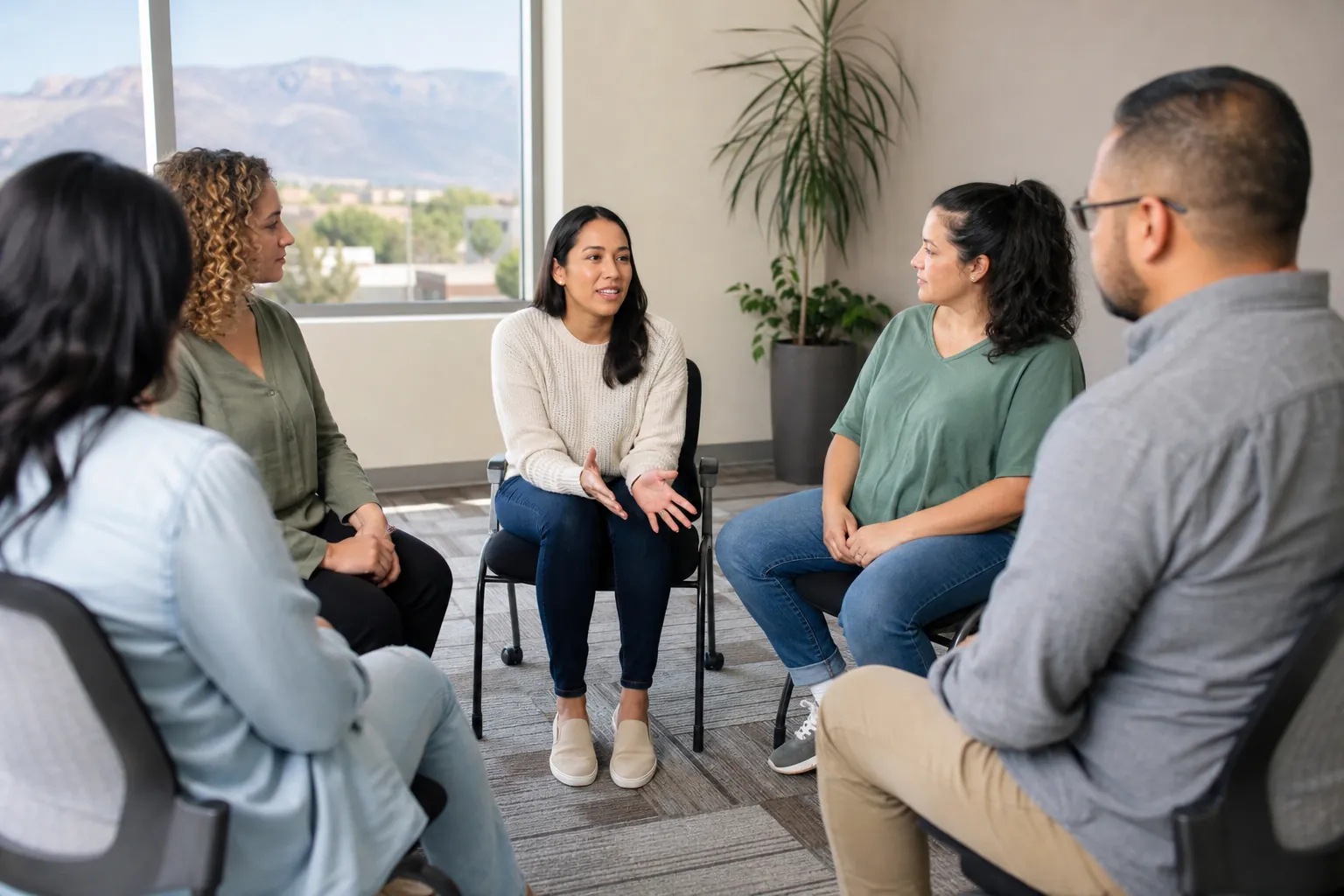 Five adults sit in a counseling circle as one woman speaks, with Albuquerque-area mountains visible through a window.