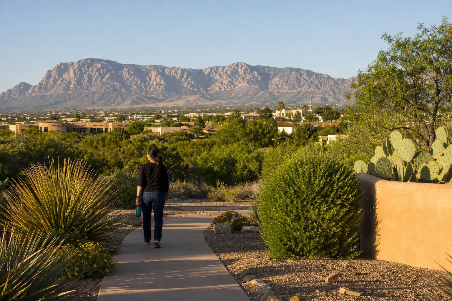 A woman walks along a paved path through desert landscaping, with low buildings and rugged mountains in the distance.
