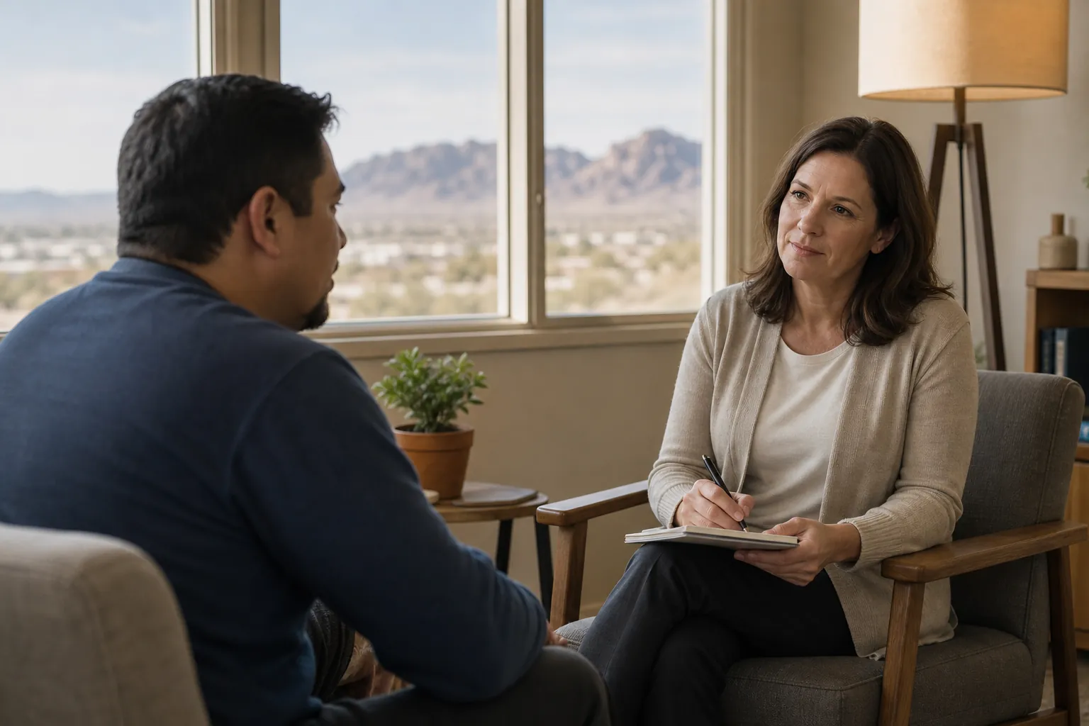 A woman holding a pen and notebook listens to a seated man in an office with desert mountains visible through the windows.