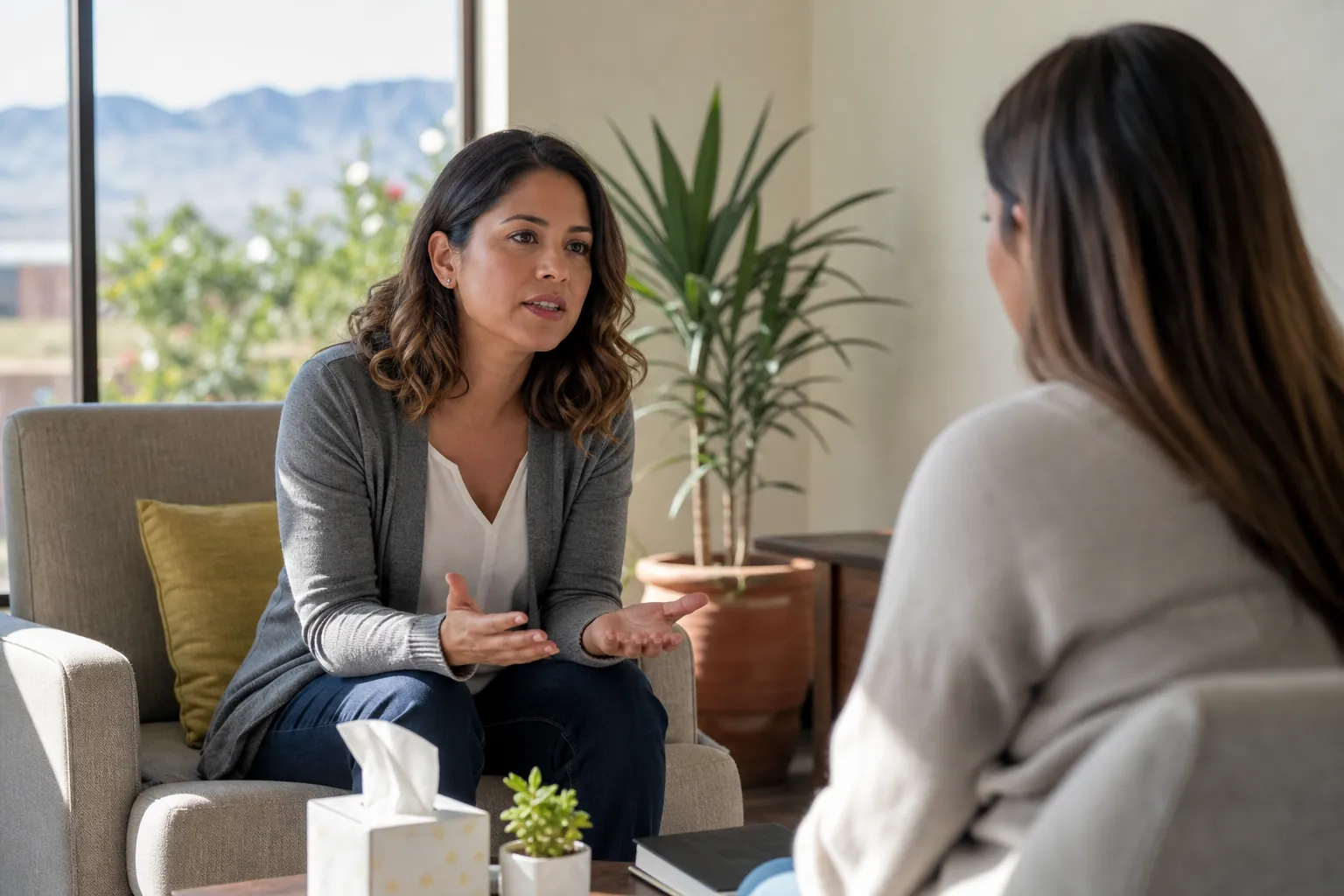 Two women sit facing each other in a bright office, one gesturing with open hands beside a table with tissues and a plant.