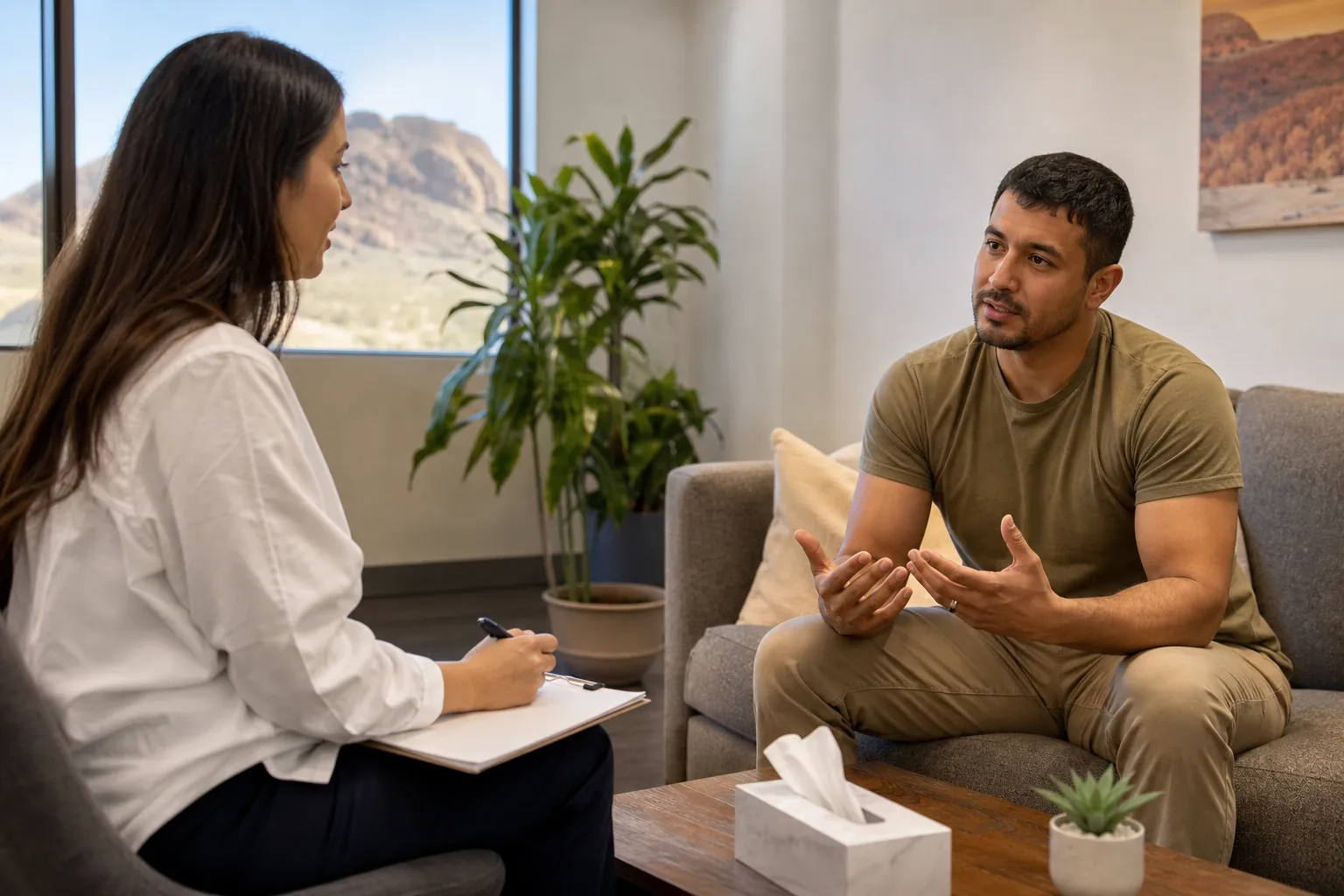 A man gestures while talking with a woman holding a pen and clipboard in an office with desert mountains outside the window.