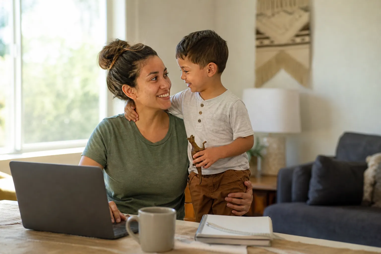 A smiling woman sits beside a laptop with a young boy holding a toy dinosaur, with a mug and notebook on the table.