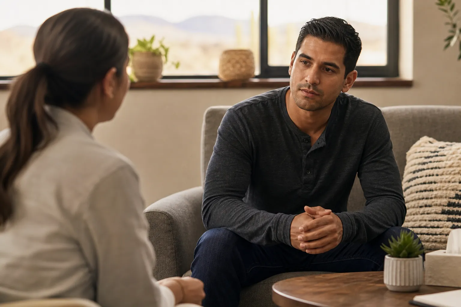 A man sits on a sofa with his hands clasped, facing a woman in a bright room with plants and a wooden coffee table.