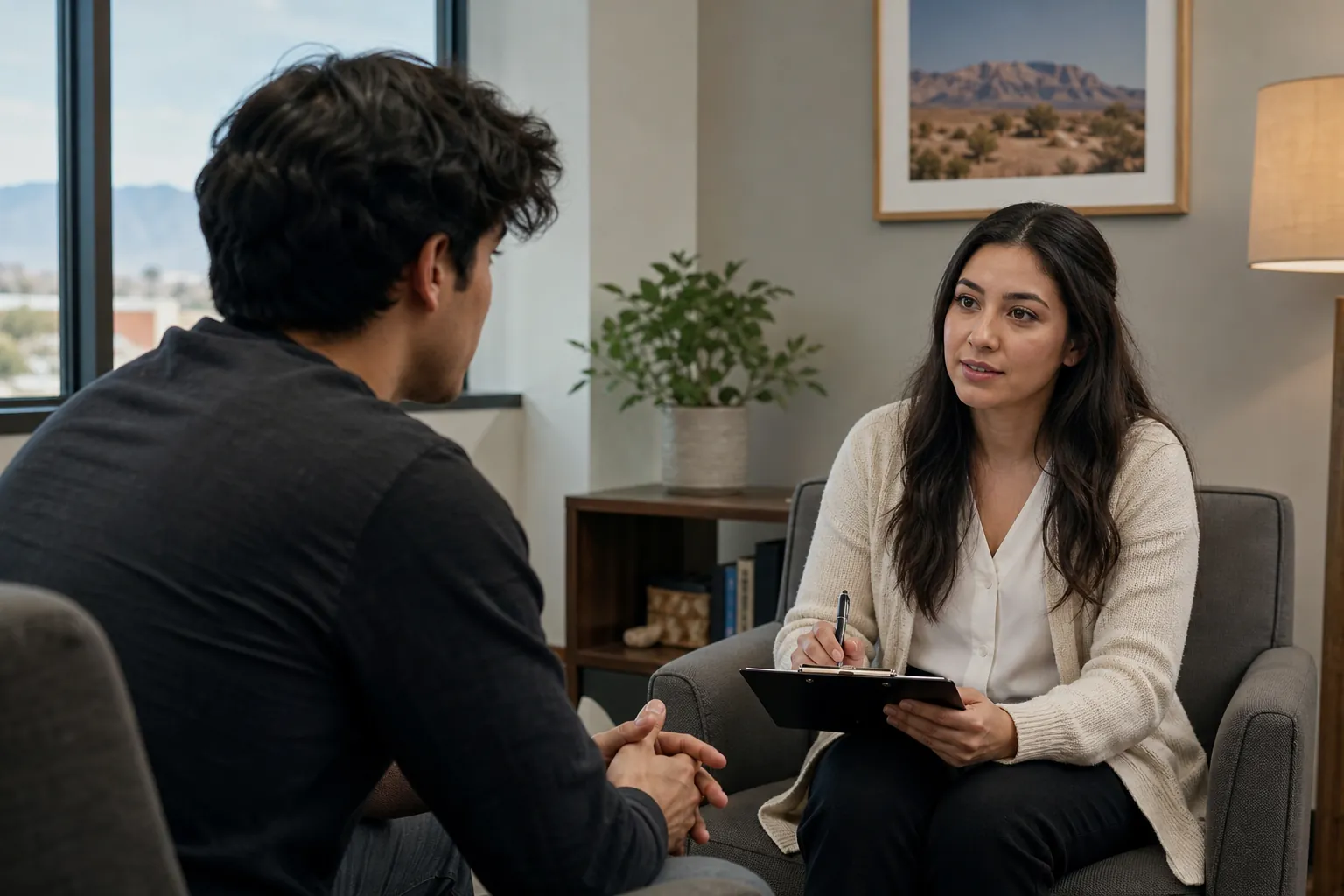A woman holding a pen and clipboard listens to a seated man in an office with a plant and framed desert landscape.