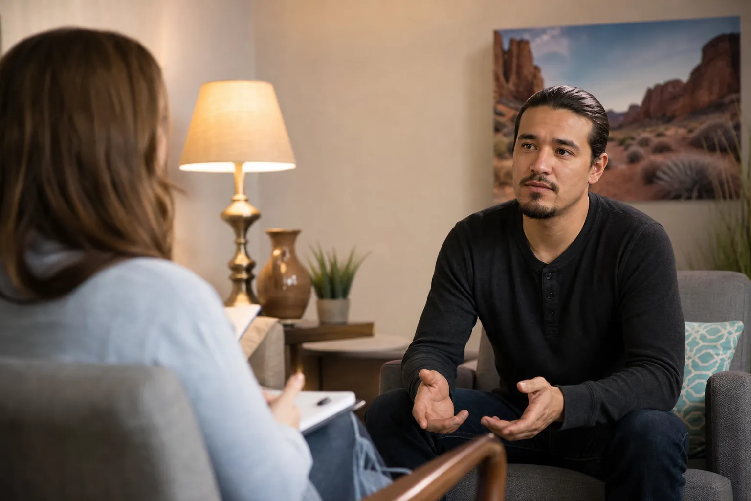 A seated man gestures while talking with a woman holding a notepad in an office with a framed desert landscape.