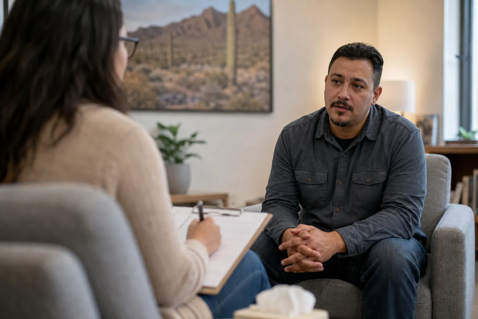 A seated man with clasped hands talks with a woman holding a pen and clipboard in an office with desert wall art.