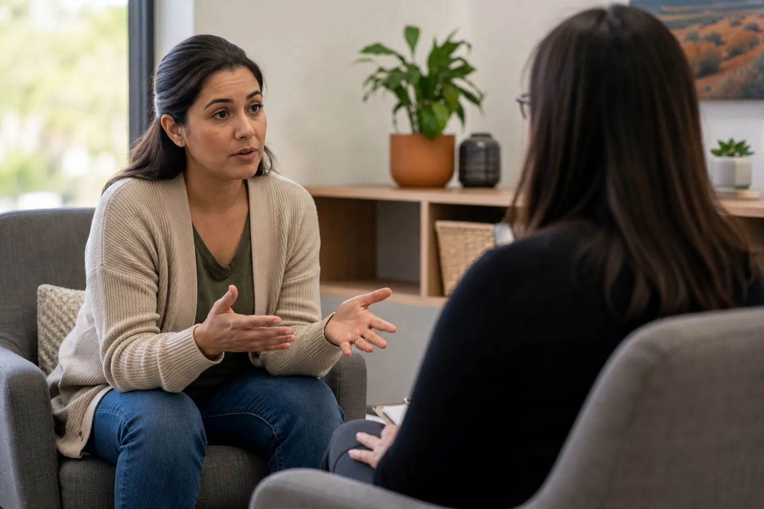 A woman gestures with both hands while talking to another woman seated opposite her in an office with plants.