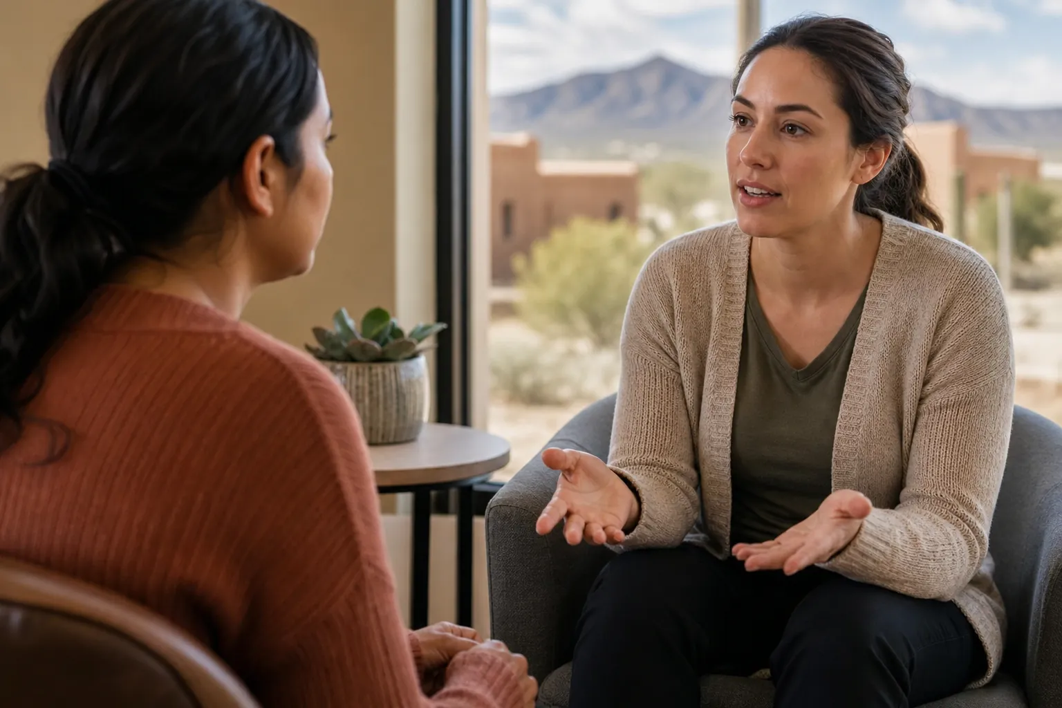 Two women sit facing each other, one speaking with open palms, beside a window overlooking mountains and adobe-style buildings.