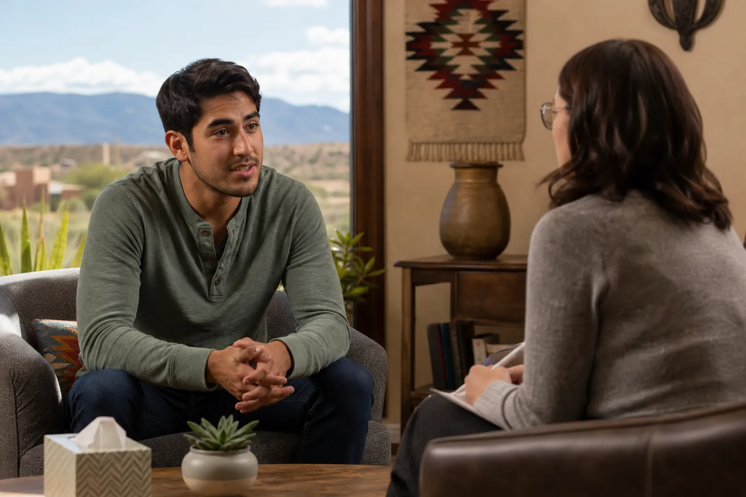 A man with clasped hands talks with a woman holding a notebook in a room with Southwestern decor and mountain views.