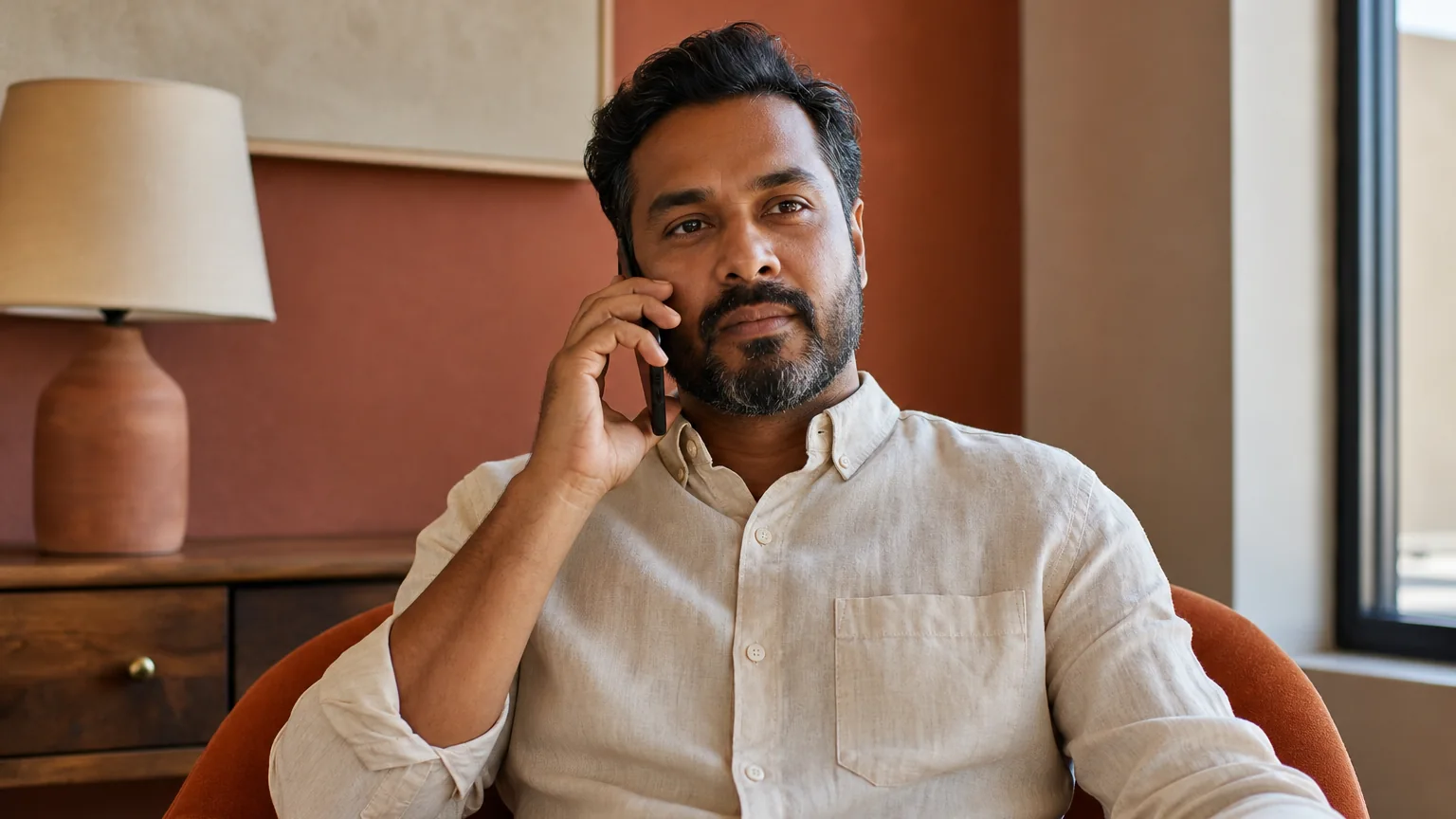 An adult man in a light button-down shirt sits in an orange armchair, holding a phone to his ear beside a window.