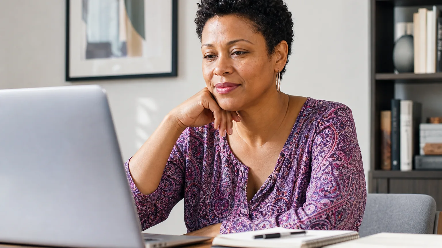 A woman in a purple patterned blouse rests her chin on her hand while looking at a laptop beside notebooks on a desk.