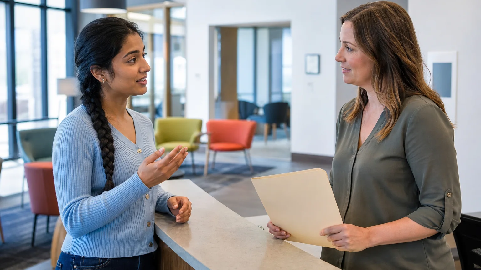 Two adult women speak across a reception counter in a bright lobby, with one gesturing and the other holding a folder.