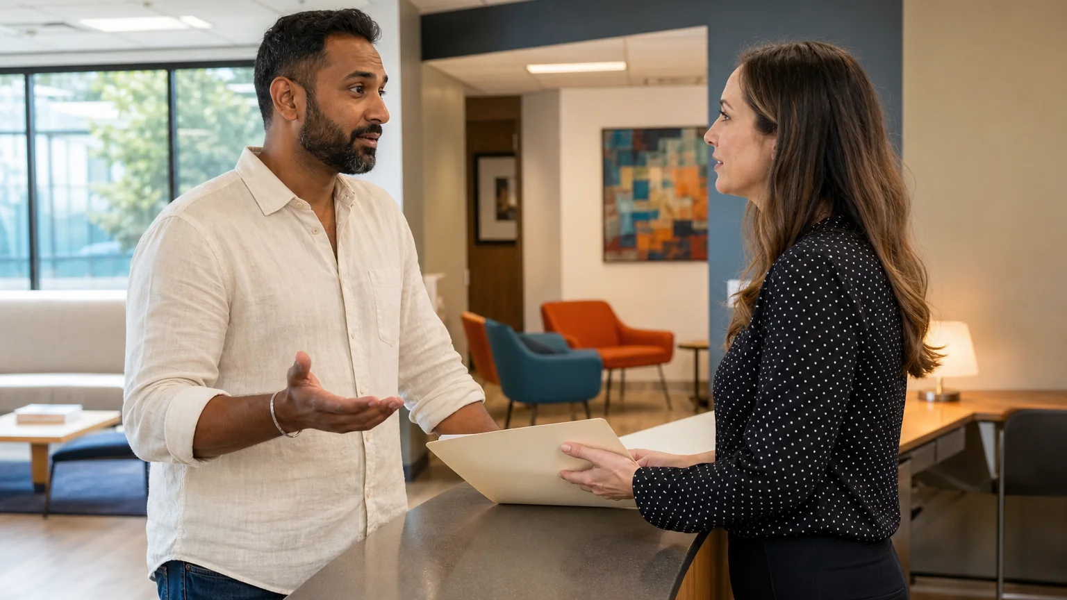 A man gestures while speaking with a woman holding a folder across a reception counter in a bright waiting area.