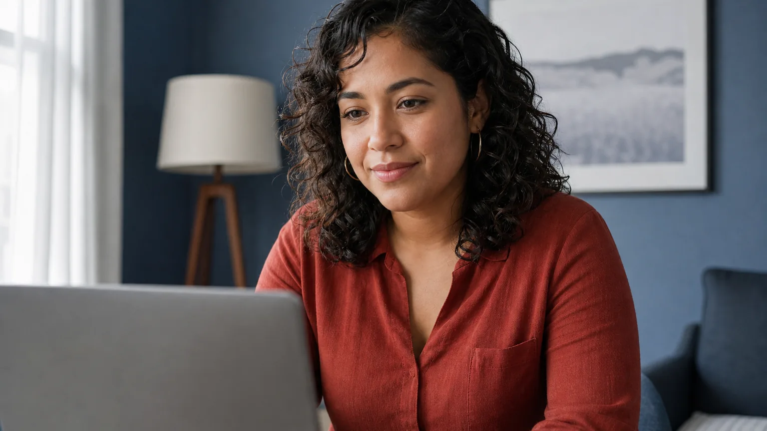 A woman in a rust-colored blouse looks at a laptop in a blue-walled room with a floor lamp and framed landscape.