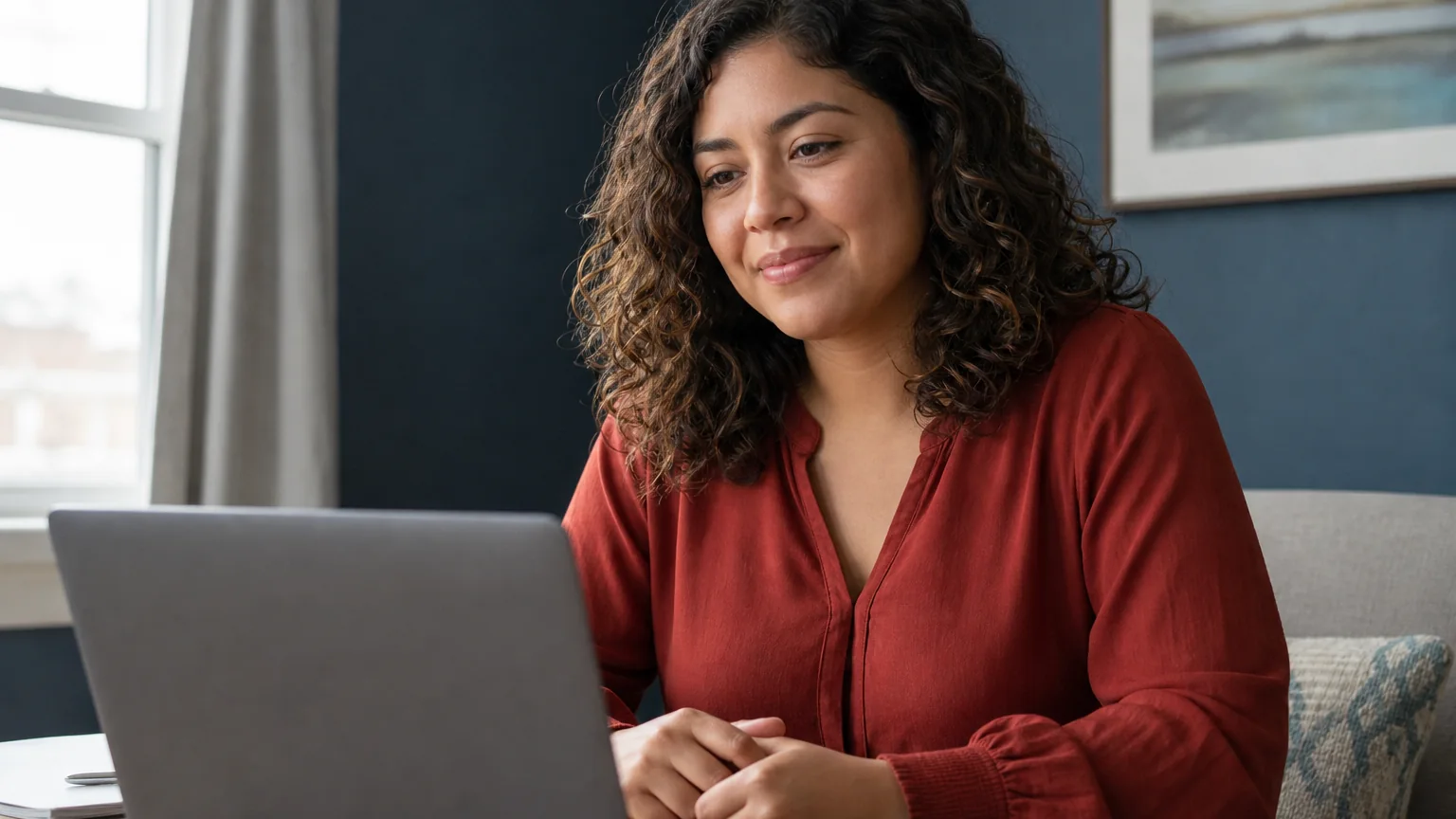 A curly-haired woman in a red blouse sits with hands clasped at a wooden table, facing a laptop in a blue-walled room.