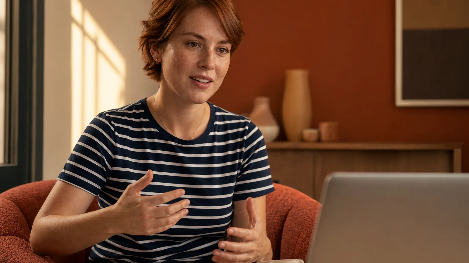 An adult woman in a striped shirt sits in a rust-colored armchair, gesturing toward an open laptop on a table.