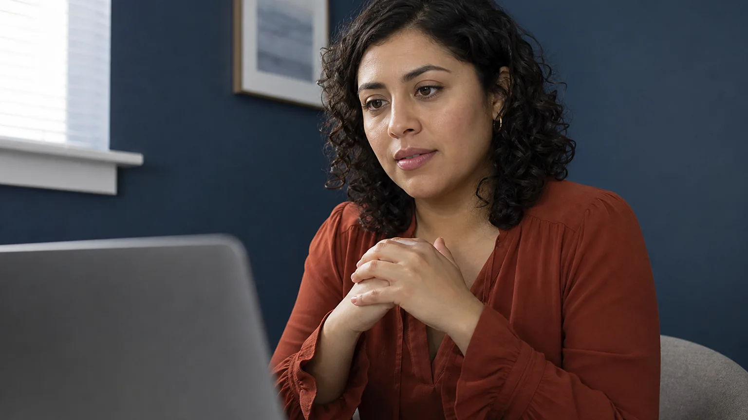A woman in a rust-colored blouse sits with her hands clasped, looking at a laptop in a blue-walled room.