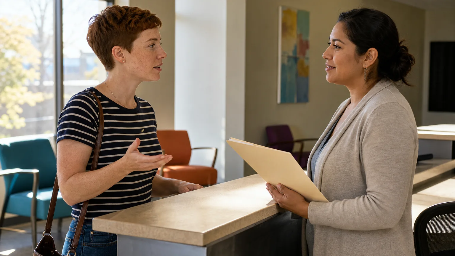 Two adults converse across a reception counter, one gesturing while the other holds a folder in a waiting area.