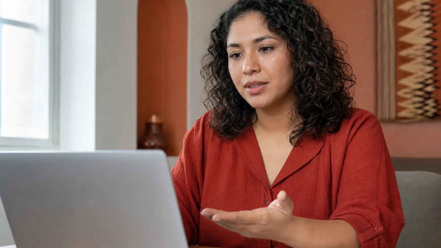 A curly-haired woman in a red blouse gestures toward an open laptop at a wooden table with a notebook beside her.