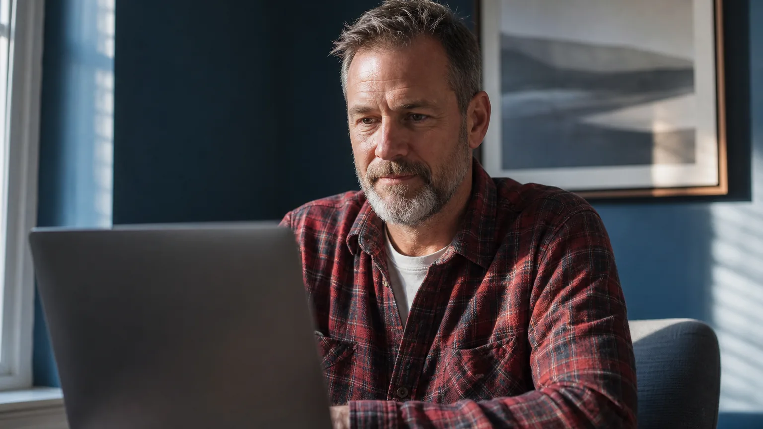 A bearded man in a red plaid shirt sits at a table using a laptop beside a window in a blue-walled room.