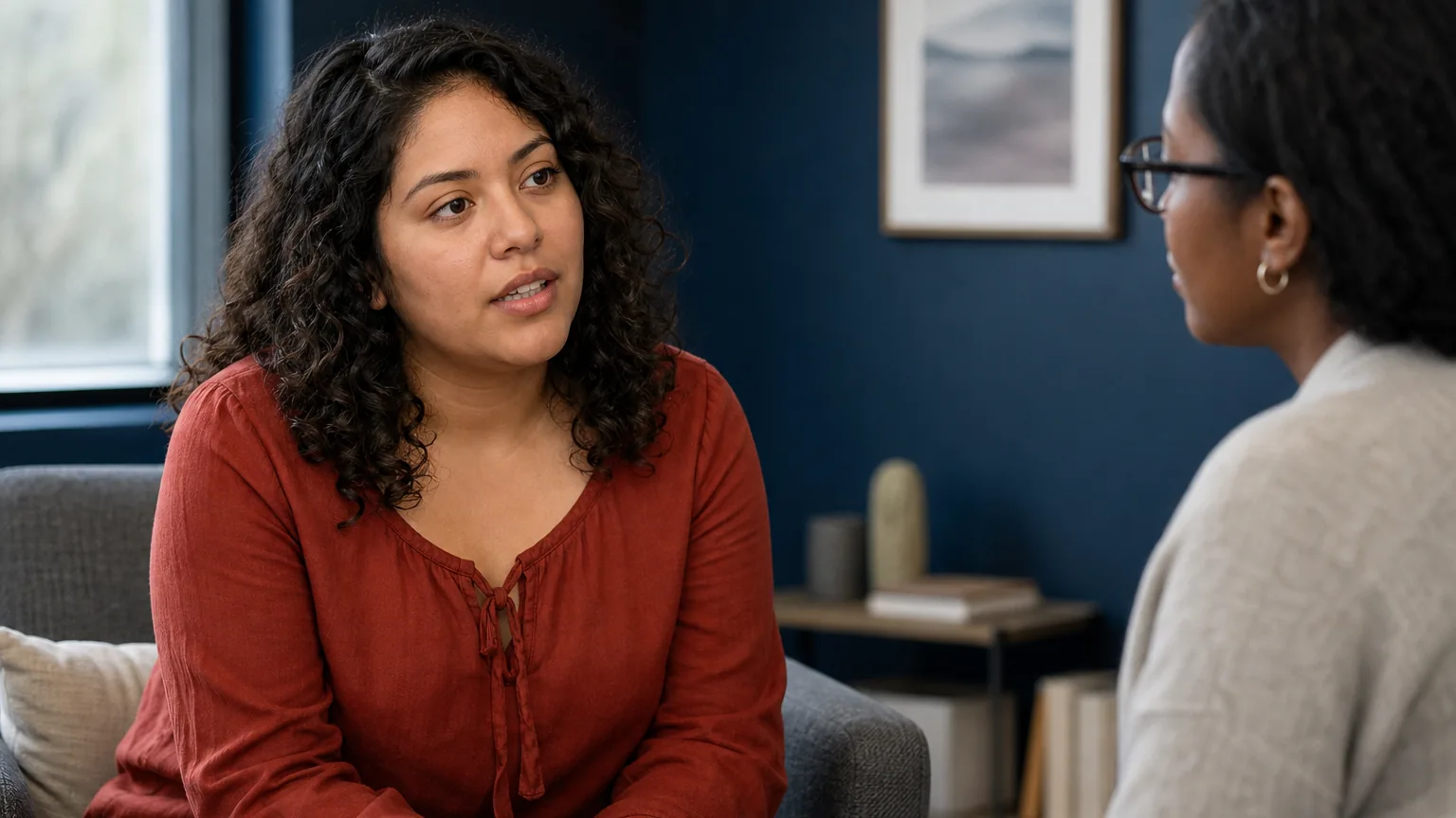 A woman in a rust-colored blouse sits on a couch, talking with a woman wearing glasses in a counseling-style office.