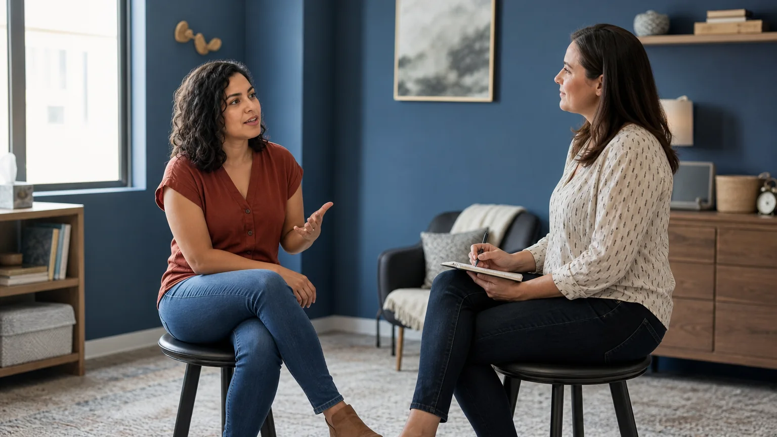 Two adult women sit facing each other in a blue-walled office, one gesturing while the other holds a notebook and pen.