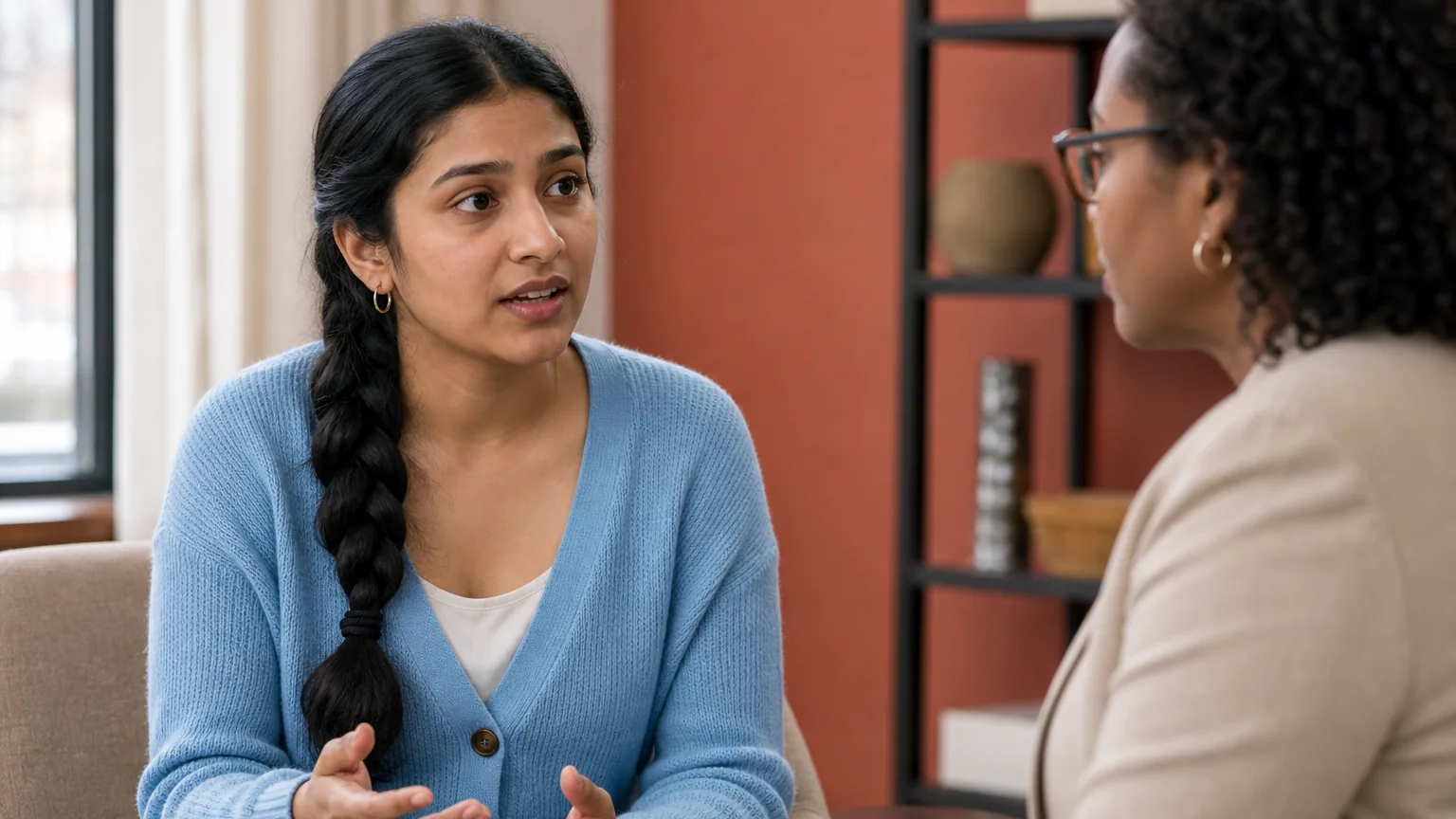 A woman in a blue cardigan gestures while speaking with a woman wearing glasses in a seated, face-to-face conversation.