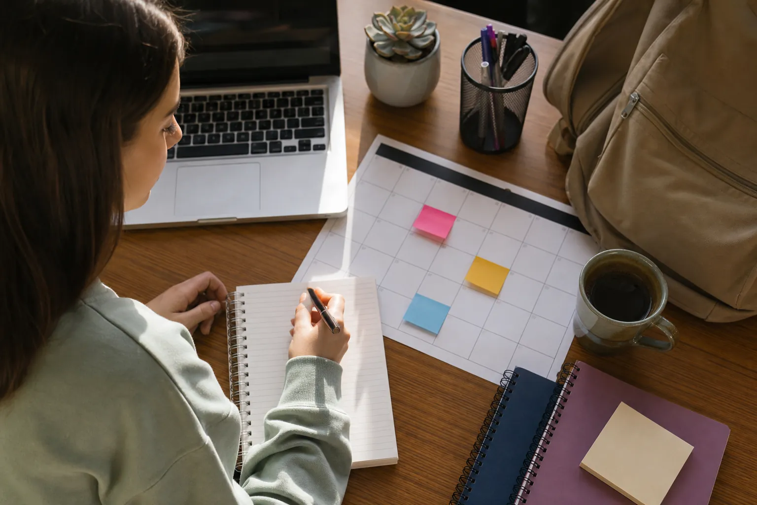 A woman holds a pen over a notebook beside a calendar with colorful sticky notes, a laptop, coffee, and a backpack.