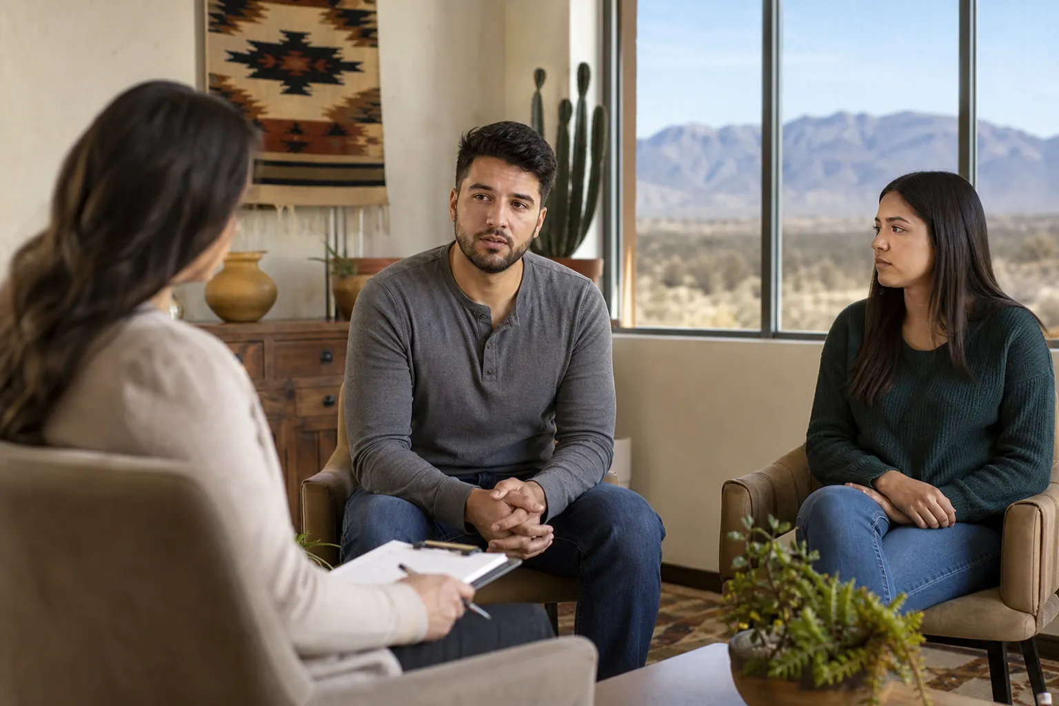 A counselor speaks with a man and woman in an office overlooking a New Mexico desert and mountain landscape.